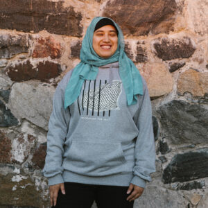 A person wearing a teal headscarf and a gray hoodie stands smiling in front of a stone wall.