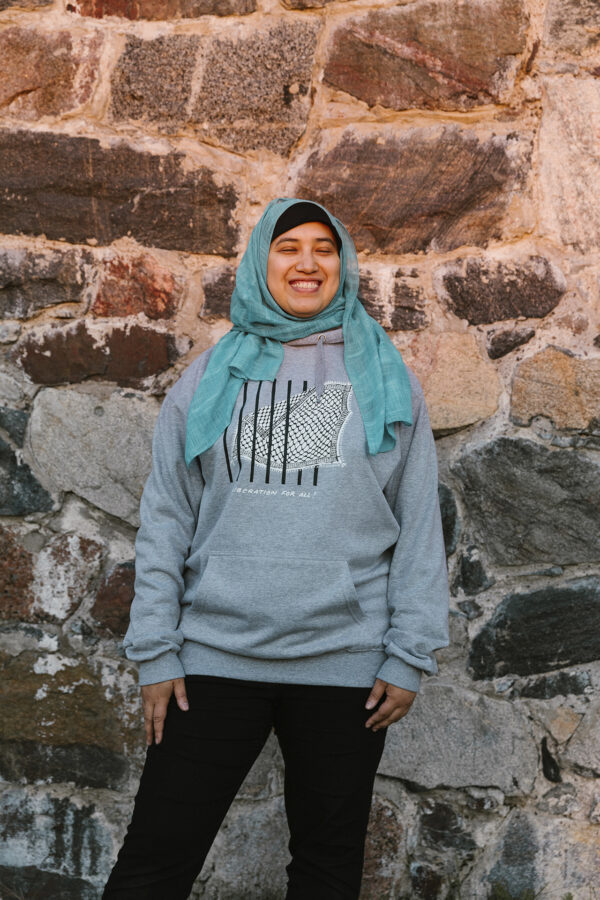 A person wearing a teal headscarf and a gray hoodie stands smiling in front of a stone wall.