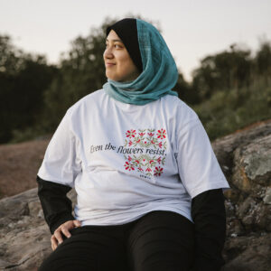 A person wearing a blue hijab and a T-shirt with floral designs sits on a rock outdoors, smiling. The shirt displays the text "Even the flowers resist." Trees and sky are visible in the background.