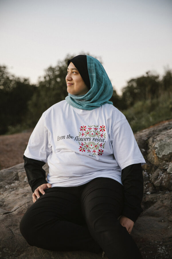 A person wearing a blue hijab and a T-shirt with floral designs sits on a rock outdoors, smiling. The shirt displays the text "Even the flowers resist." Trees and sky are visible in the background.