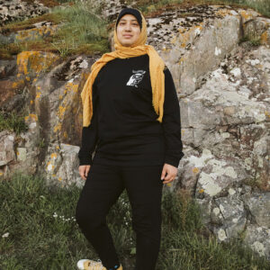 A person wearing a black outfit and a yellow headscarf stands on grassy rocks, looking at the camera. They have yellow sneakers, and the background features rocky terrain with patches of greenery and moss.