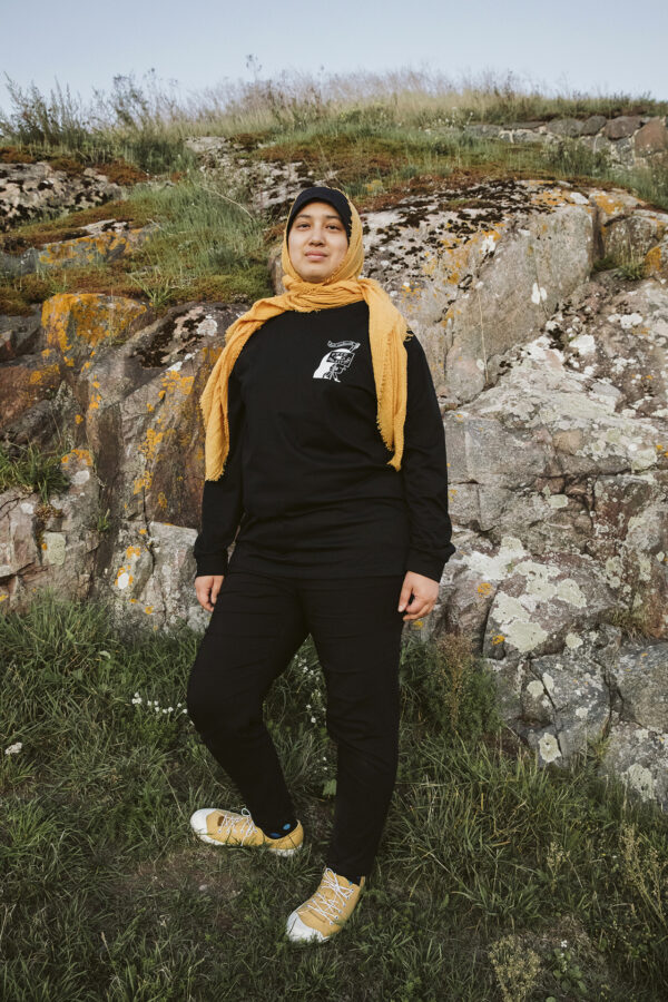 A person wearing a black outfit and a yellow headscarf stands on grassy rocks, looking at the camera. They have yellow sneakers, and the background features rocky terrain with patches of greenery and moss.