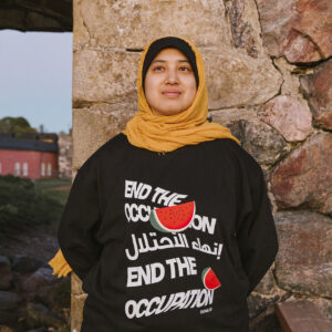 A person wearing a hijab and a black sweater with the text "End the Occupation" and a watermelon design stands in front of a stone wall, with grassy landscape and a building visible in the background.