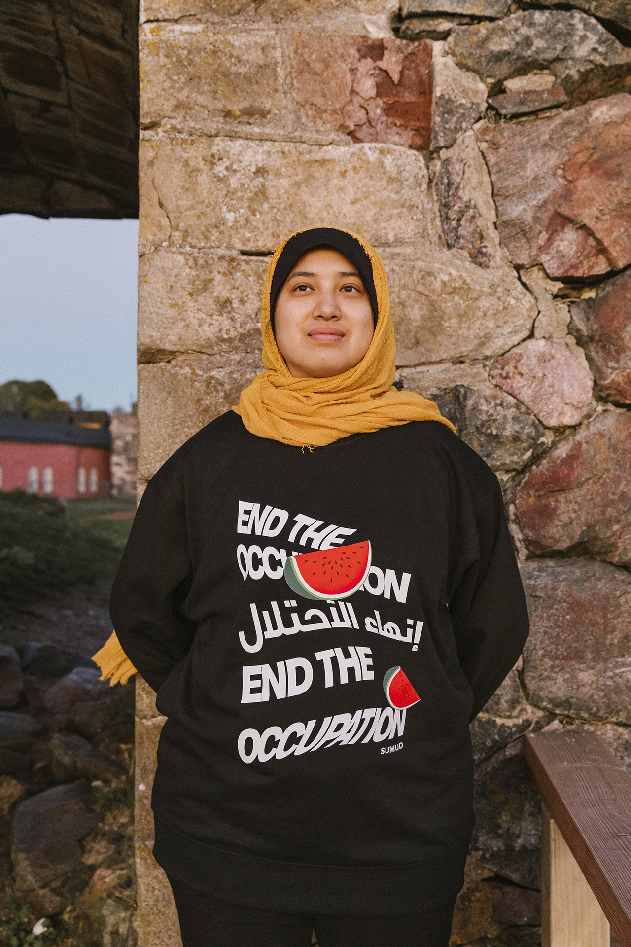 A person wearing a hijab and a black sweater with the text "End the Occupation" and a watermelon design stands in front of a stone wall, with grassy landscape and a building visible in the background.