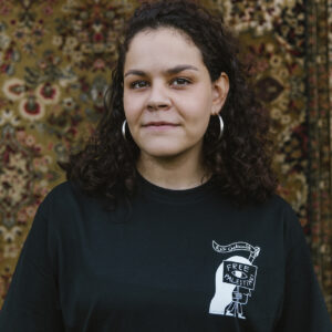 A person with curly hair and hoop earrings stands in front of a patterned rug, wearing a black T-shirt with a "Free Palestine" design. They are looking at the camera with a neutral expression.