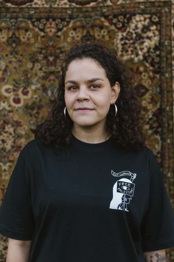 A person with curly hair and hoop earrings stands in front of a patterned rug, wearing a black T-shirt with a "Free Palestine" design. They are looking at the camera with a neutral expression.