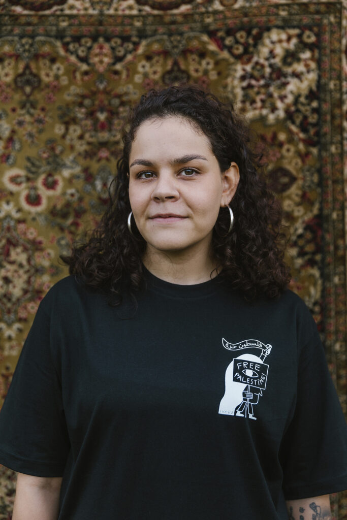 A person with curly hair and hoop earrings stands in front of a patterned rug, wearing a black T-shirt with a "Free Palestine" design. They are looking at the camera with a neutral expression. 