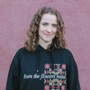 A person with curly hair standing against a textured pink wall, wearing a black hoodie with a floral design and the text "Even the flowers resist" on the front.