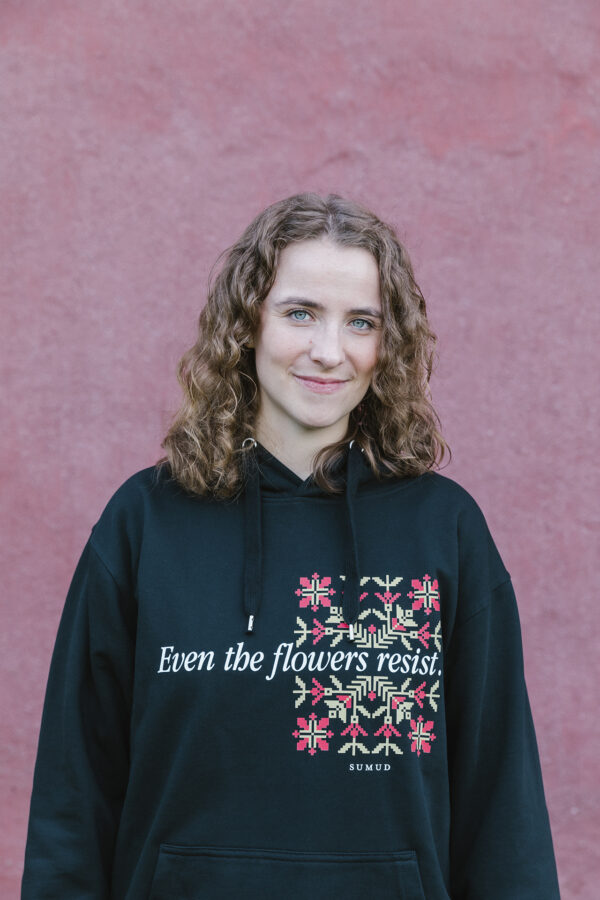 A person with curly hair standing against a textured pink wall, wearing a black hoodie with a floral design and the text "Even the flowers resist" on the front.