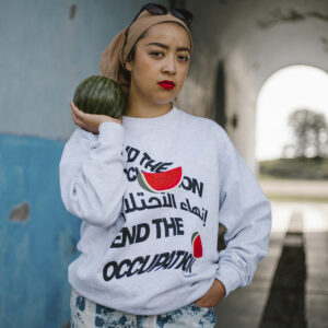 A person stands in a hallway with a squashes| vegetableses on their shoulder, wearing a grey sweatshirt with a bold statement and a watermelon graphic. They have a beige headscarf, sunglasses on top, and are in blue patterned jeans.