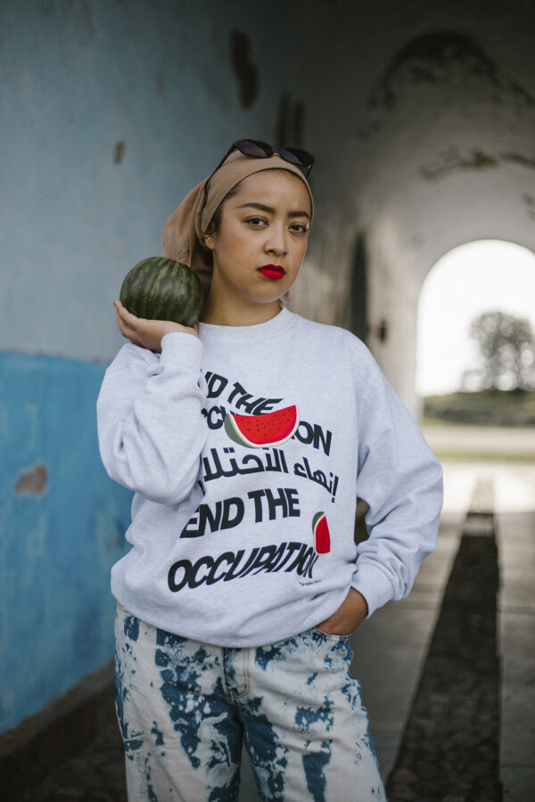 A person stands in a hallway with a squashes| vegetableses on their shoulder, wearing a grey sweatshirt with a bold statement and a watermelon graphic. They have a beige headscarf, sunglasses on top, and are in blue patterned jeans.