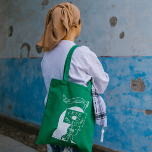 A person wearing a beige headscarf and a white long-sleeve shirt stands with their back to the camera. They carry a green tote bag with "Free Palestine" written on it. The background is a blue and gray textured wall.