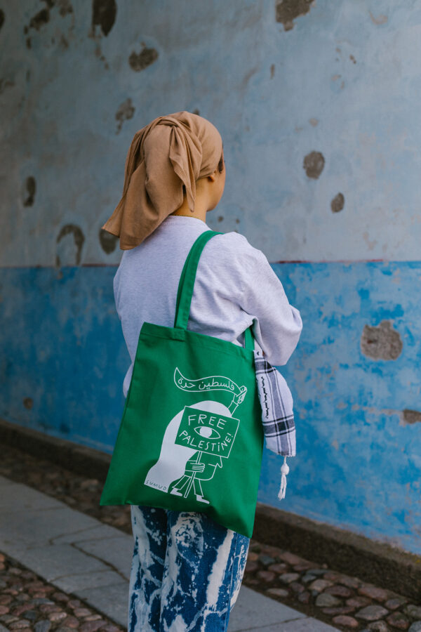 A person wearing a beige headscarf and a white long-sleeve shirt stands with their back to the camera. They carry a green tote bag with "Free Palestine" written on it. The background is a blue and gray textured wall.