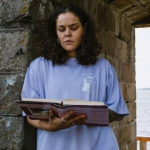 A person with curly hair stands by a stone archway, wearing a light blue t-shirt, reading a book. The background shows a glimpse of a body of water and the sky.