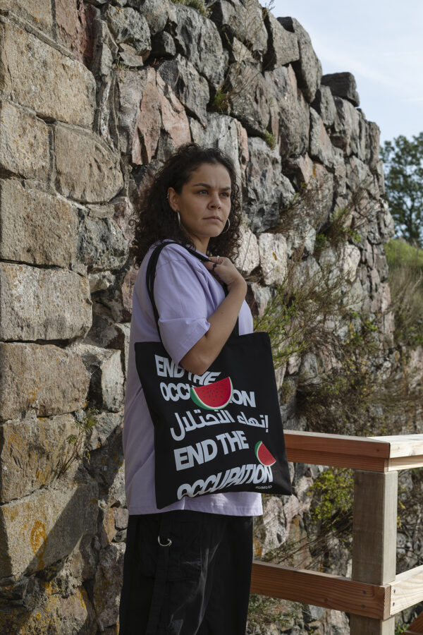 A person with curly hair wearing a purple shirt stands by a stone wall, holding a black tote bag that reads "End the Occupation" in English and Arabic, with a red graphic. There are wooden and metal railings nearby and green foliage in the background.