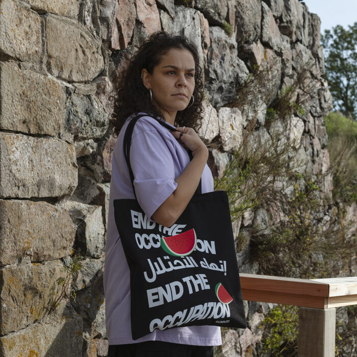 A person with curly hair wearing a purple shirt stands by a stone wall, holding a black tote bag that reads "End the Occupation" in English and Arabic, with a red graphic. There are wooden and metal railings nearby and green foliage in the background.