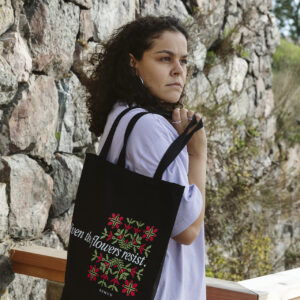 A person with curly hair stands outdoors next to a stone wall, holding a black tote bag with colorful flower patterns and text. The person wears a light purple shirt and looks thoughtfully into the distance.