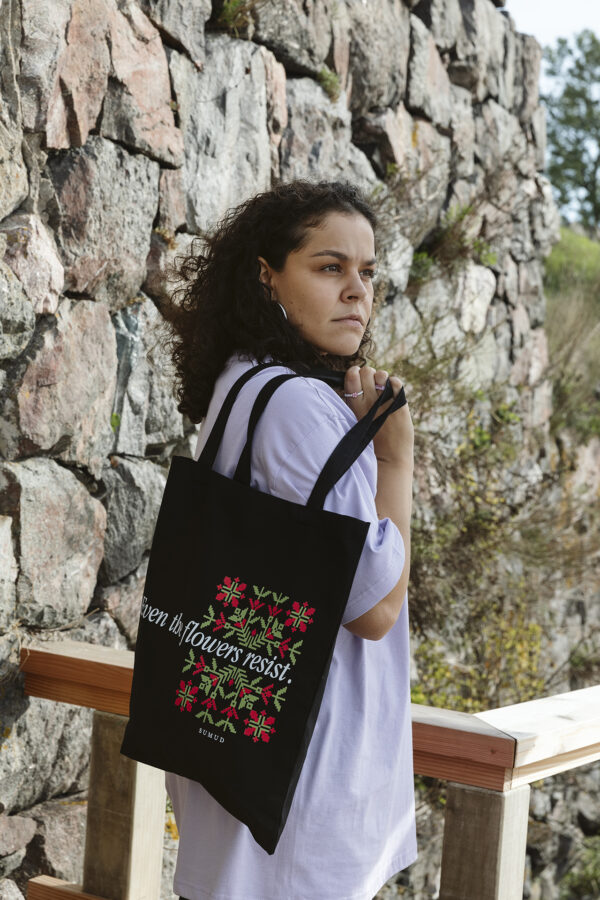 A person with curly hair stands outdoors next to a stone wall, holding a black tote bag with colorful flower patterns and text. The person wears a light purple shirt and looks thoughtfully into the distance.