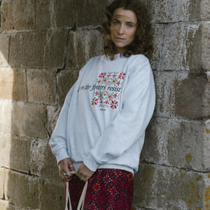 A woman with curly hair stands against a stone wall, wearing a light gray sweatshirt with floral designs and text. She holds a woven bag and wears a long, dark red skirt with a pattern. The setting is rustic and outdoors.