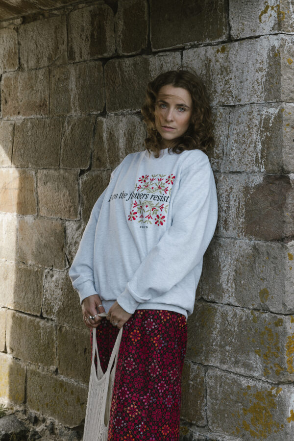 A woman with curly hair stands against a stone wall, wearing a light gray sweatshirt with floral designs and text. She holds a woven bag and wears a long, dark red skirt with a pattern. The setting is rustic and outdoors.