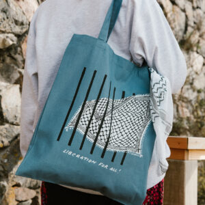 A person with long hair, wearing a gray sweatshirt and a patterned skirt, carries a blue tote bag featuring a barbed wire design and the text "LIBERATION FOR ALL!" The person stands in front of a stone wall.