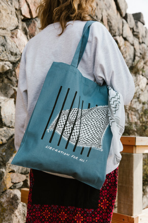 A person with long hair, wearing a gray sweatshirt and a patterned skirt, carries a blue tote bag featuring a barbed wire design and the text "LIBERATION FOR ALL!" The person stands in front of a stone wall.