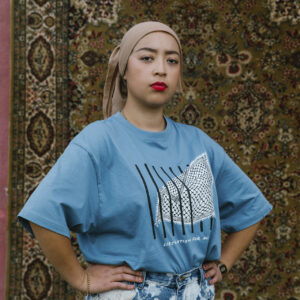 A person with bright red lipstick stands confidently with hands on hips, wearing a blue T-shirt and patterned jeans in front of a detailed patterned rug.