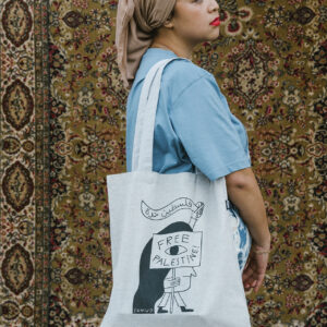 A person in a blue shirt and headscarf holds a tote bag that reads "Free Palestine" against a patterned rug backdrop. The tote bag also features an illustrated character.