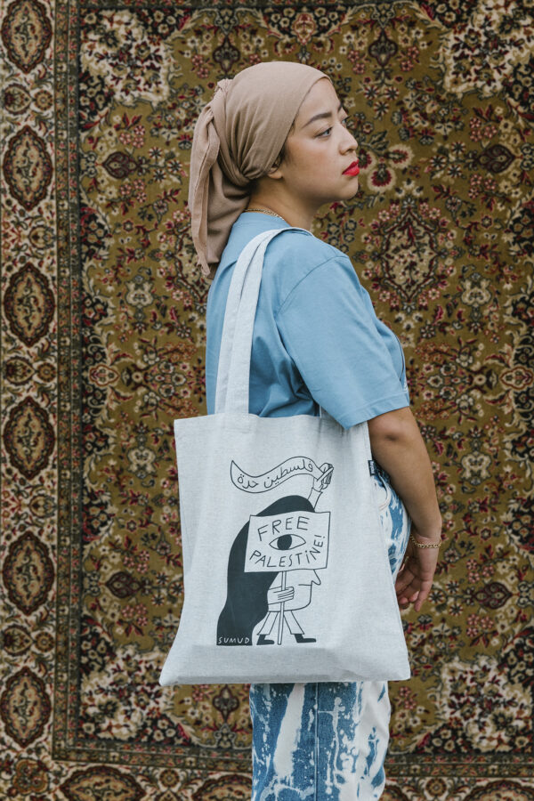 A person in a blue shirt and headscarf holds a tote bag that reads "Free Palestine" against a patterned rug backdrop. The tote bag also features an illustrated character.