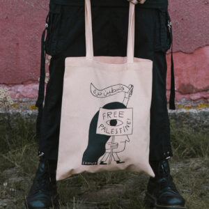 A person holds a beige tote bag with a graphic design. The design features a stylized woman in a headscarf holding a banner that reads "Free Palestine" in English and Arabic. The person is wearing black clothing and standing on grass in front of a pink wall.