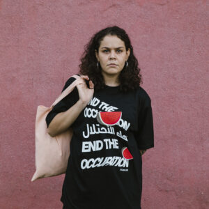 A person with curly hair stands in front of a pink wall, wearing a black T-shirt with white text and red watermelon graphics that say, “End the Occupation” in multiple languages. They carry a light-colored tote bag.