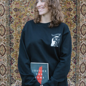 A smiling person with curly hair stands in front of a patterned rug, holding a book titled "Justice for Some." They are wearing a black sweatshirt with a design on it and light blue jeans.