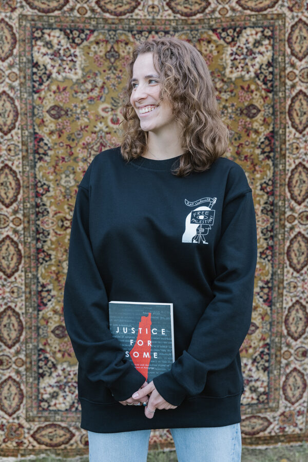 A smiling person with curly hair stands in front of a patterned rug, holding a book titled "Justice for Some." They are wearing a black sweatshirt with a design on it and light blue jeans.