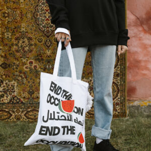 A person wearing a black sweater and light blue jeans holds a white tote bag with text that reads "END THE OCCUPATION" in English and Arabic, with watermelon graphics. They stand on grass in front of a patterned rug and pink wall.