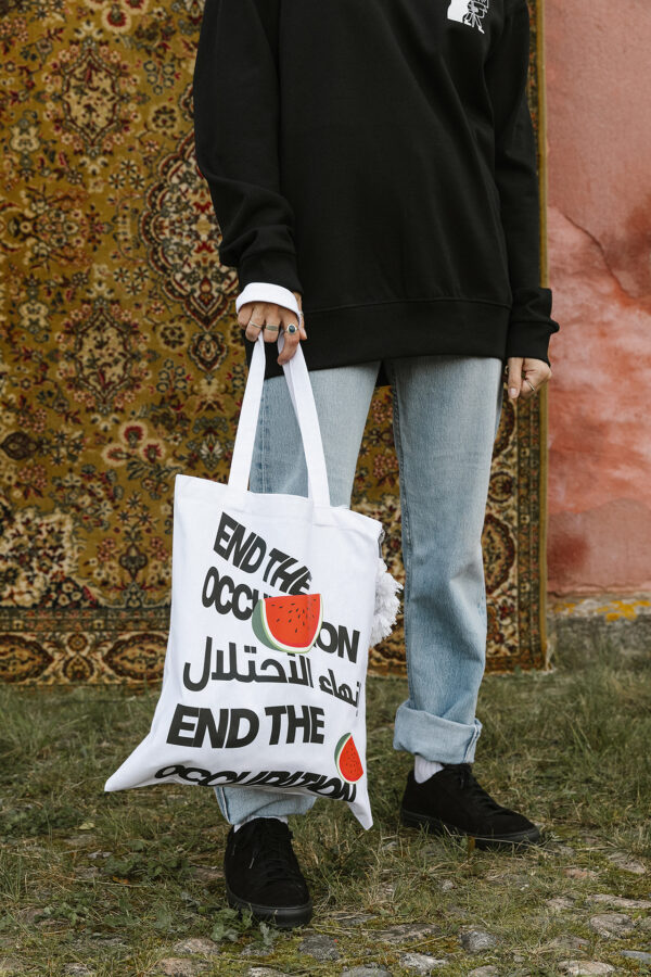 A person wearing a black sweater and light blue jeans holds a white tote bag with text that reads "END THE OCCUPATION" in English and Arabic, with watermelon graphics. They stand on grass in front of a patterned rug and pink wall.