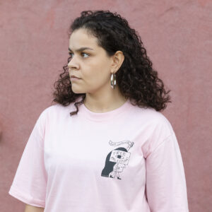 A person with curly hair stands in front of a pink wall, wearing a light pink T-shirt with an illustrated design. They have hoop earrings and visible tattoos on their arm, looking to the side with a neutral expression.