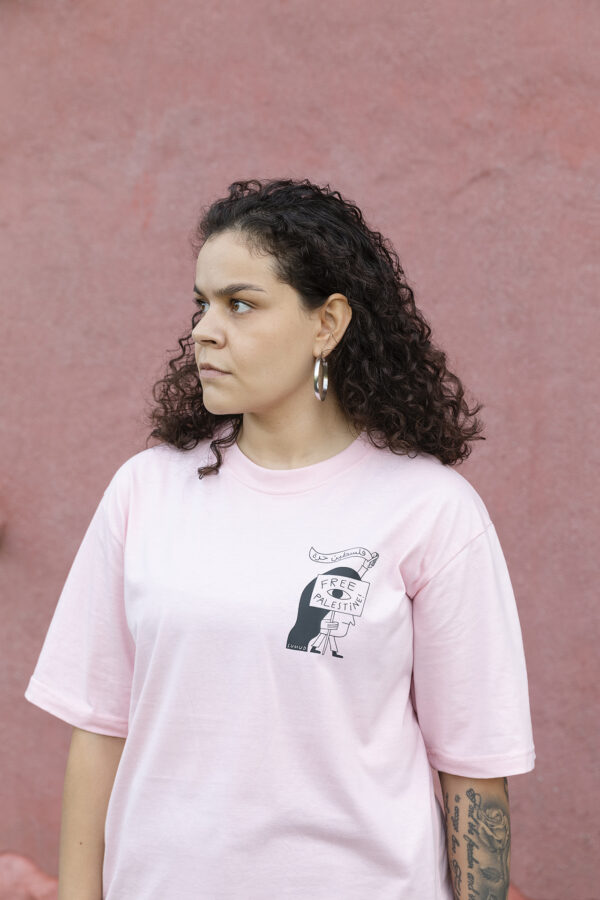 A person with curly hair stands in front of a pink wall, wearing a light pink T-shirt with an illustrated design. They have hoop earrings and visible tattoos on their arm, looking to the side with a neutral expression.