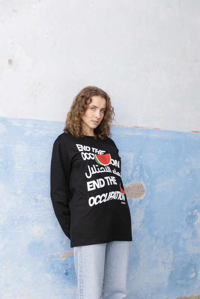 A person with curly hair stands against a two-tone wall, wearing a black long-sleeve shirt with the text "End the Occupation" and a red and green graphic. They have a neutral expression and are looking at the camera.