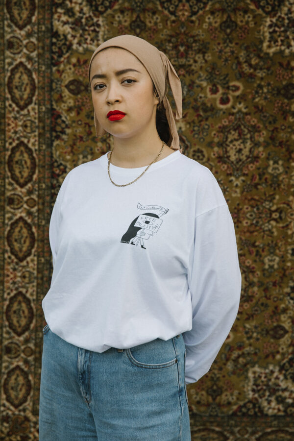 A person stands in front of an ornate tapestry, wearing a beige headscarf, white long-sleeve shirt with a small graphic, blue jeans, and red lipstick. Their hands are behind their back, and they look directly at the camera.