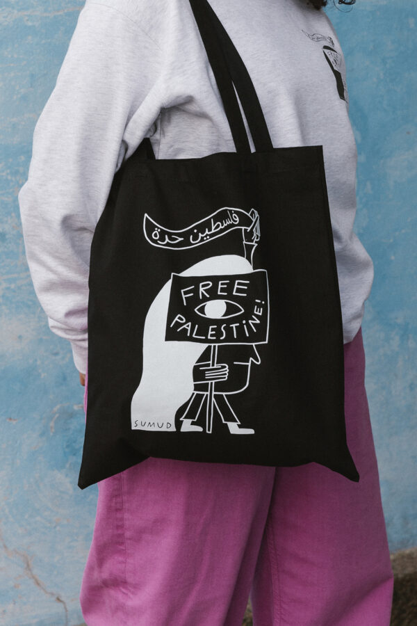 A person stands against a textured blue wall wearing a gray sweater and purple pants. They are holding a black tote bag with a graphic that reads "Free Palestine" in English and Arabic, featuring a stylized character.