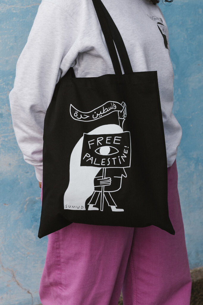 A person stands against a textured blue wall wearing a gray sweater and purple pants. They are holding a black tote bag with a graphic that reads "Free Palestine" in English and Arabic, featuring a stylized character. 
