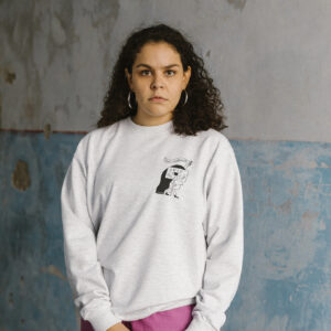 A person with long curly hair stands against a textured wall. They wear a light gray sweatshirt with a graphic on the chest and hold a black strap. The wall is weathered with patches of blue and gray.