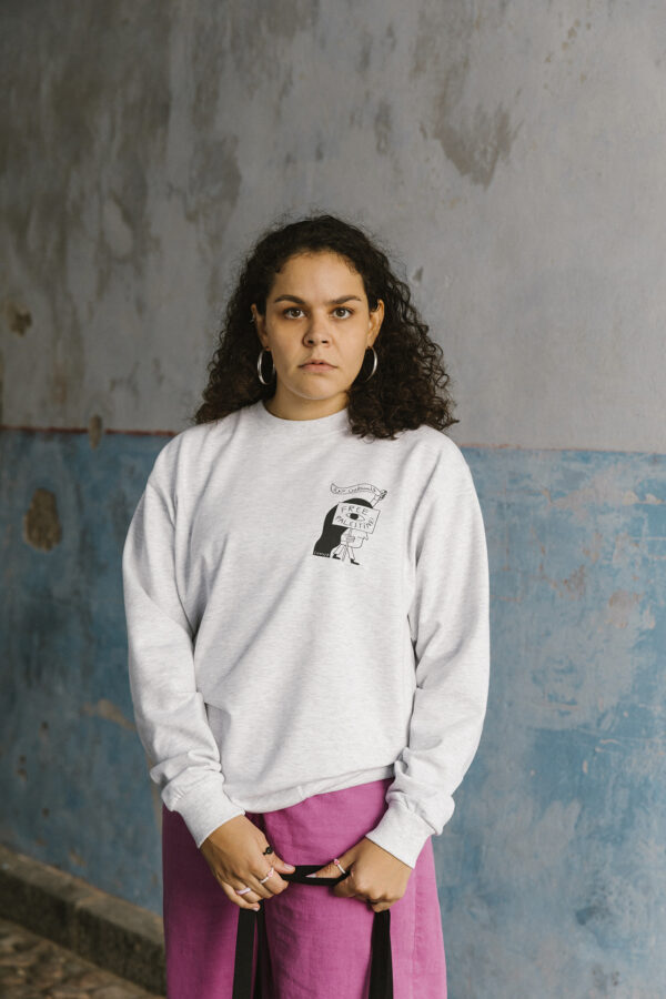 A person with long curly hair stands against a textured wall. They wear a light gray sweatshirt with a graphic on the chest and hold a black strap. The wall is weathered with patches of blue and gray.