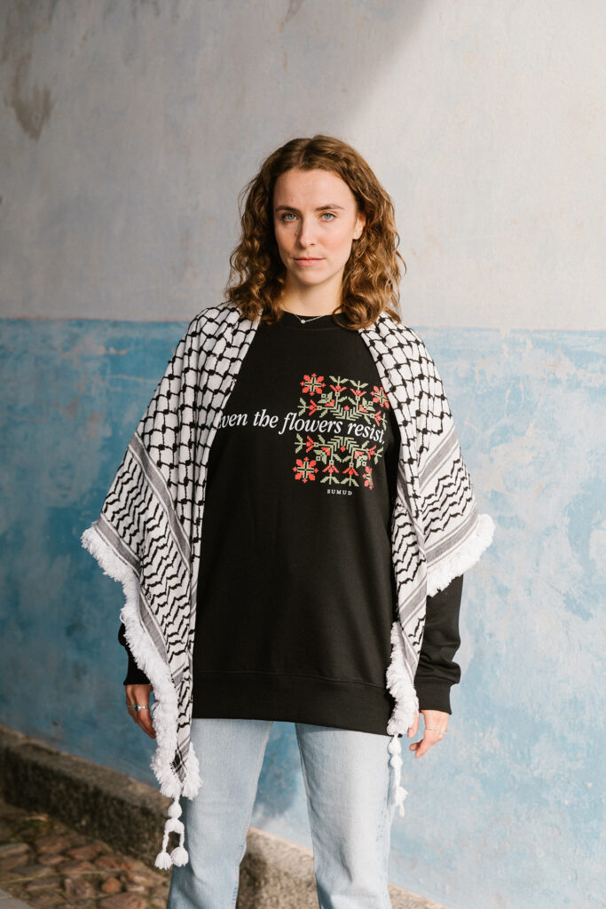 A woman with curly hair stands against a blue wall, wearing a black sweater with floral design and text, and a checkered scarf draped over her shoulders. She is also wearing light blue jeans. 