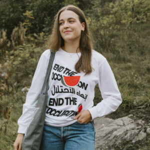 A woman stands outdoors wearing a white long-sleeve shirt with "End the Occupation" in multiple languages and a watermelon design. She carries a gray bag and is in a grassy area with trees in the background.