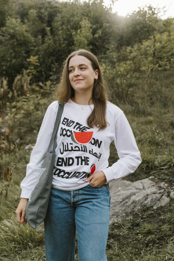 A woman stands outdoors wearing a white long-sleeve shirt with "End the Occupation" in multiple languages and a watermelon design. She carries a gray bag and is in a grassy area with trees in the background.