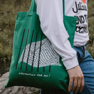 A person wearing a white shirt and blue jeans holds a green tote bag with a net design and the words "LIBERATION FOR ALL!" printed on it. The background is grassy and rocky terrain.