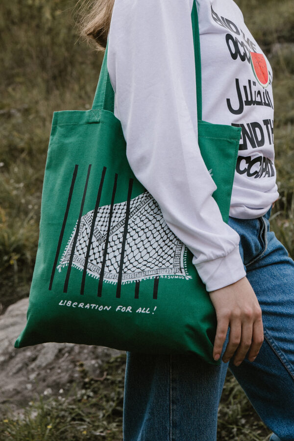 A person wearing a white shirt and blue jeans holds a green tote bag with a net design and the words "LIBERATION FOR ALL!" printed on it. The background is grassy and rocky terrain.