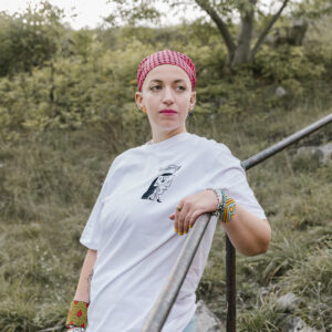 A person with short hair wearing a red and white checkered headscarf and white T-shirt leans on a metal railing outdoors. They have colorful bracelets and stand in a grassy area with trees in the background.