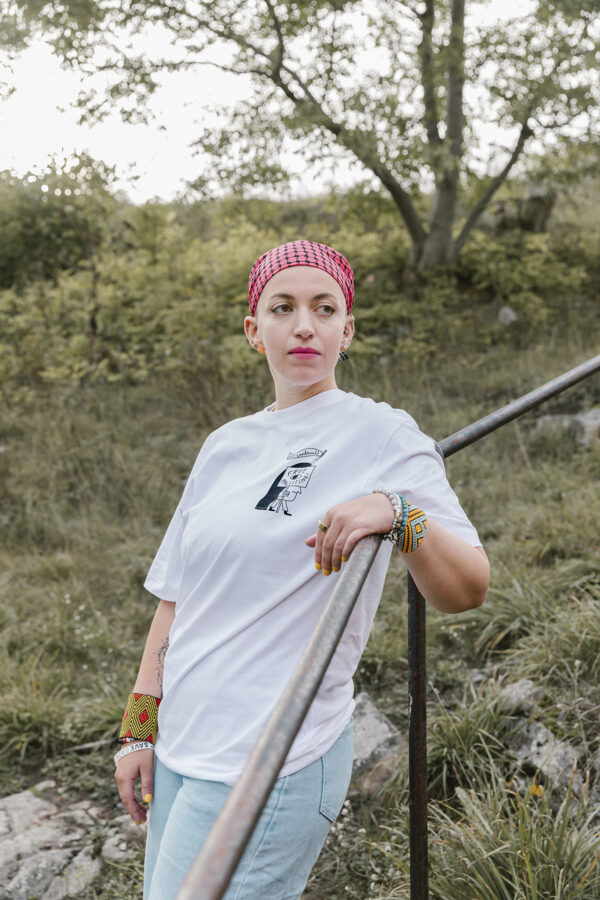 A person with short hair wearing a red and white checkered headscarf and white T-shirt leans on a metal railing outdoors. They have colorful bracelets and stand in a grassy area with trees in the background.